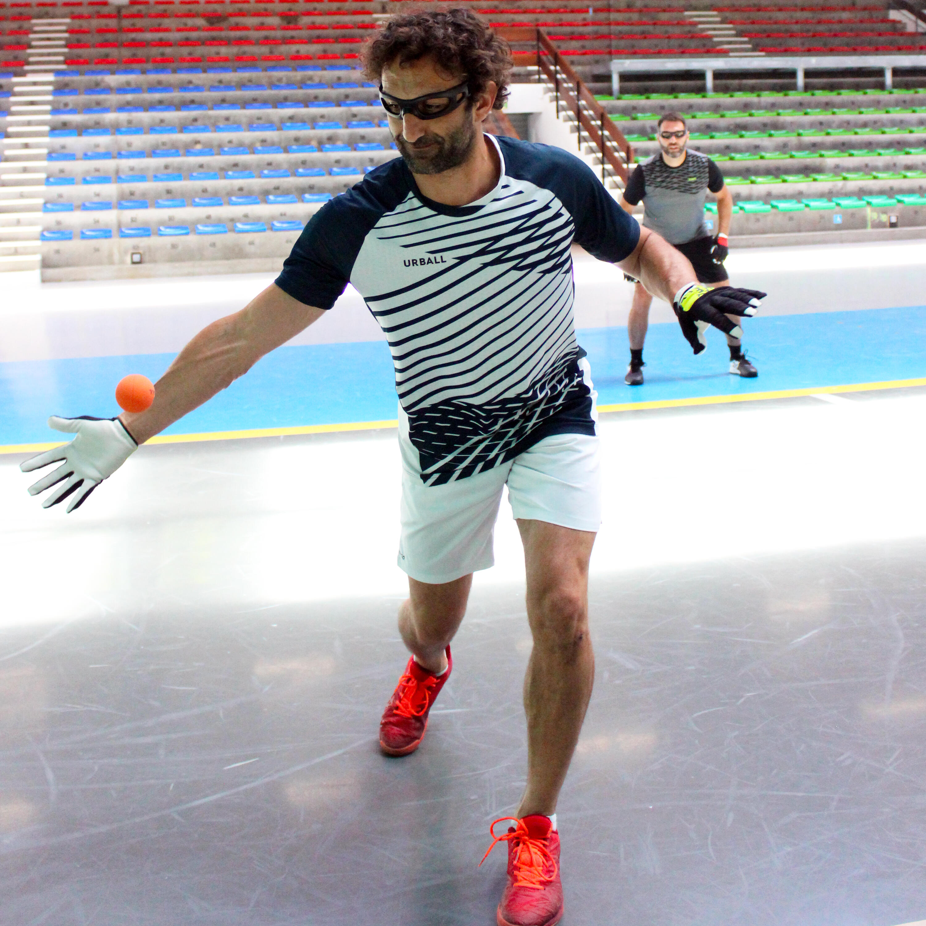 Pelota-Spieler in Aktion mit Handschuhen und Schutzbrille. Nahaufnahme des Sportoutfits (blau-weiß gestreift, weiße Shorts) und der roten Turnschuhe auf dem Spielfeld.