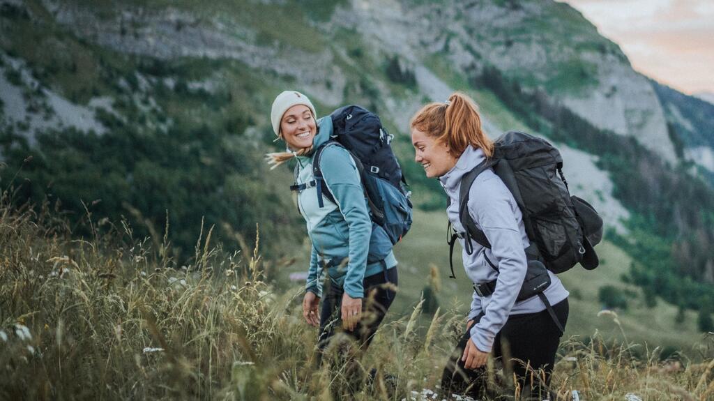 Zwei Frauen mit Rucksäcken wandern durch grüne Berglandschaft, hohe Gräser und sanfte Hügel im Hintergrund.