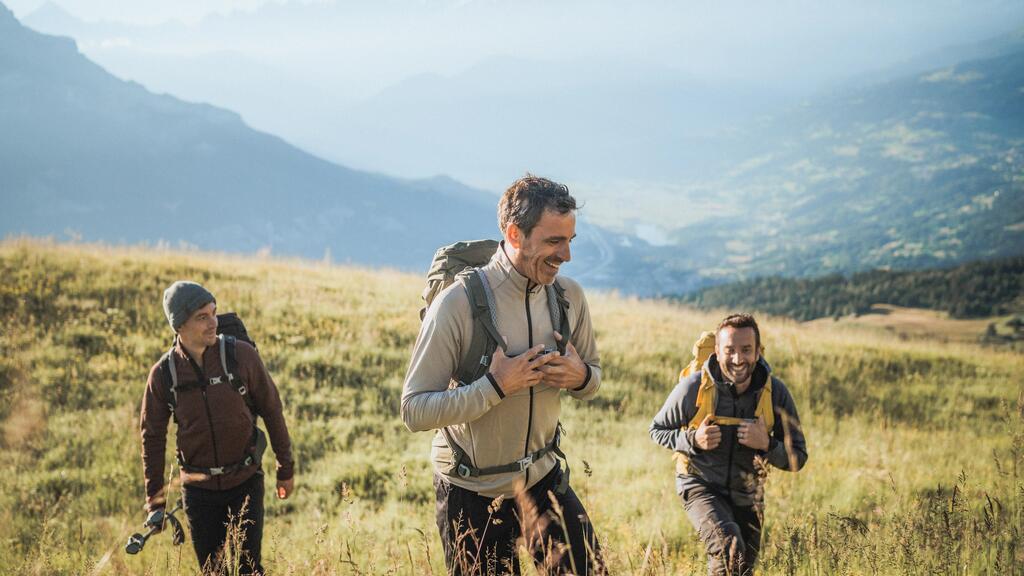 Drei lachende Männer mit Wanderrucksäcken wandern bei Sonnenschein über eine weite, goldene Bergwiese.