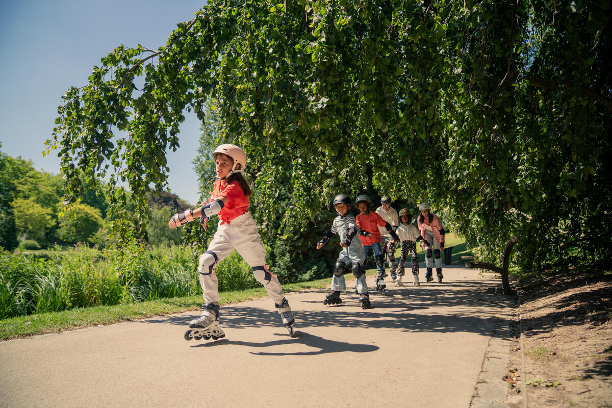 Grupa dece i odraslih sa kacigama i kompletnom zaštitnom opremom veselo vozi rolere po sunčanoj stazi u parku ispod drveća.