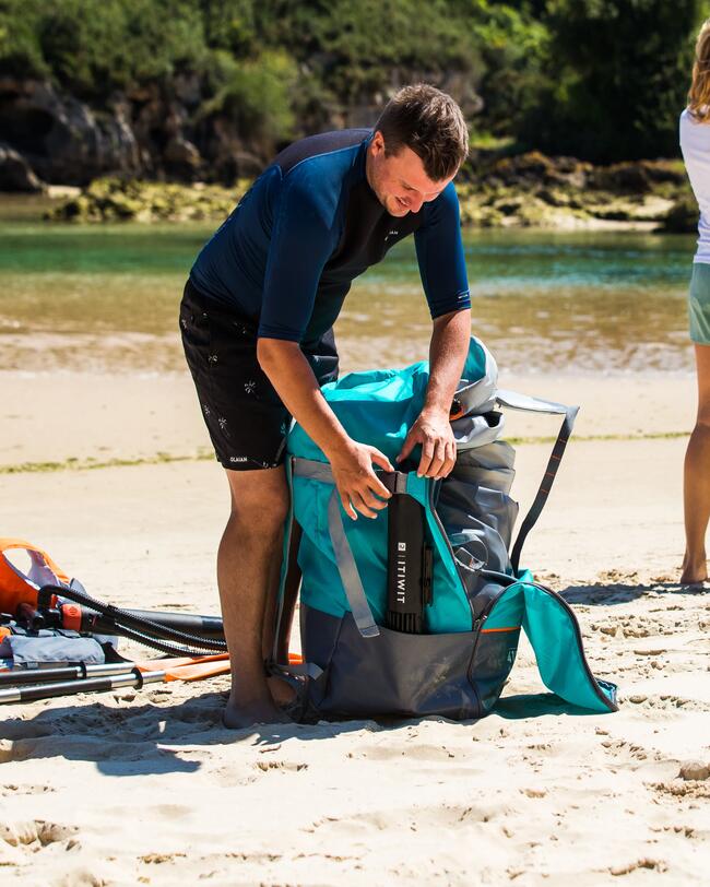 Mann am Strand, packt türkis-schwarzen Rucksack, sonniges Wetter, sandiger Boden, grüne Pflanzen im Hintergrund.