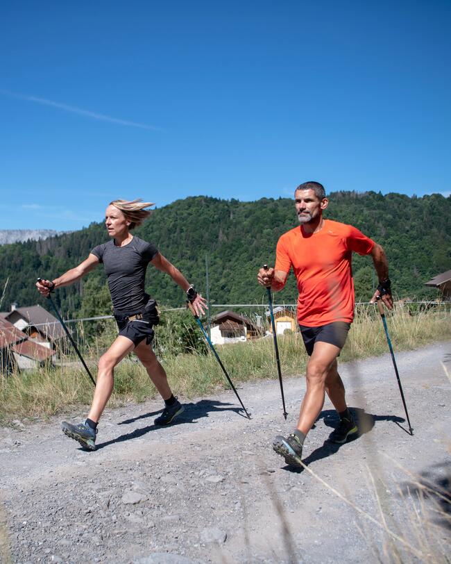 Zwei Menschen beim Nordic Walking auf einem Schotterweg, umgeben von grünen Hügeln unter klarem, blauem Himmel.