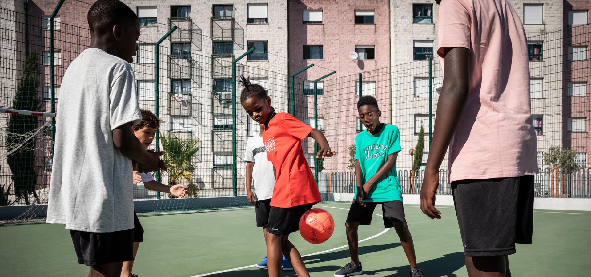 Crianças a fazer acrobacias com bola de futsal numa quadra no meio de prédios