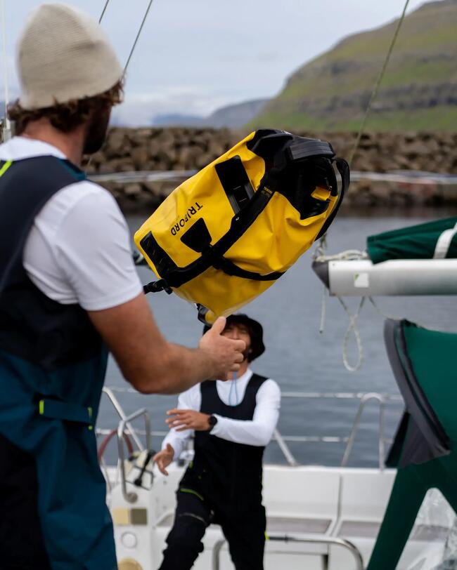Zwei Personen auf einem Boot, werfen gelbe Tasche. Tragen Segelkleidung, Hintergrund aus Wasser und Hügeln.