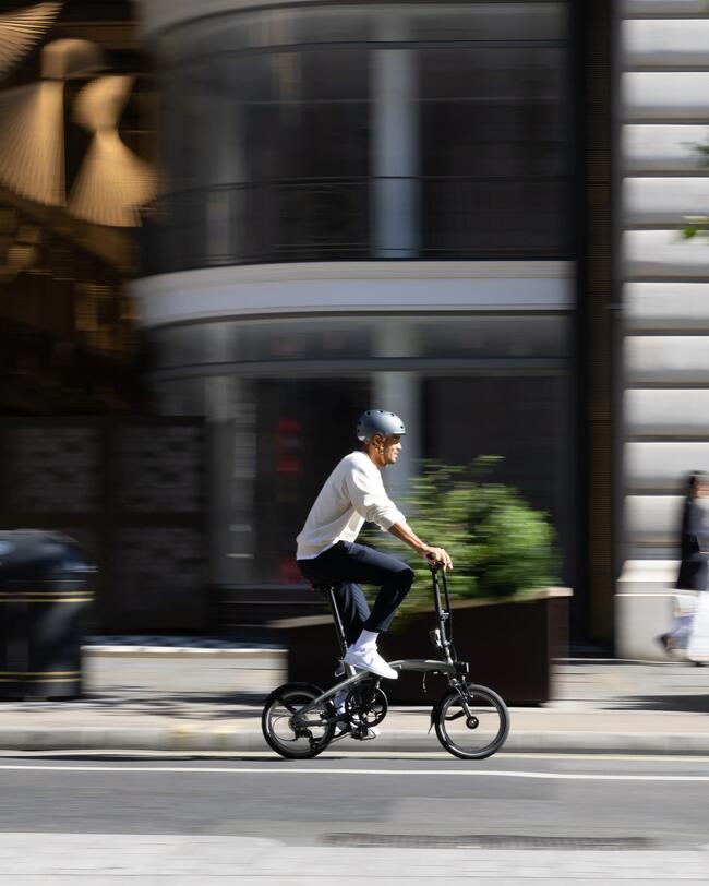 Mann mit Helm fährt auf Klapprad eine Straße entlang; im Hintergrund Stadtszene unscharf