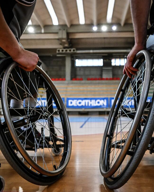 Zwei Personen in Basketballrollstühlen, silberne Räder, Holzboden, Sporthalle im Hintergrund.