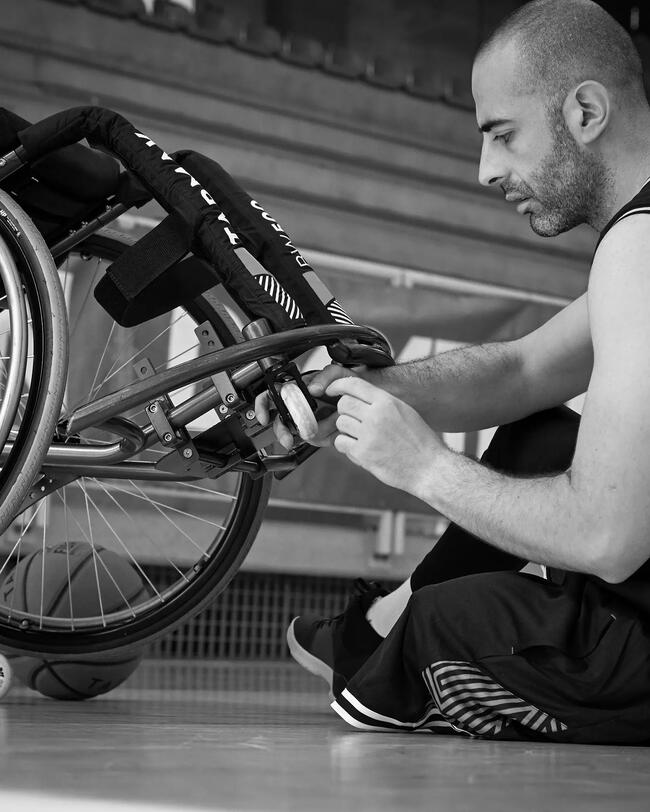 Mann passt Basketballrollstuhl an, schwarz-weiß, glatte Texturen, runde Formen, parkettierter Hallenboden.