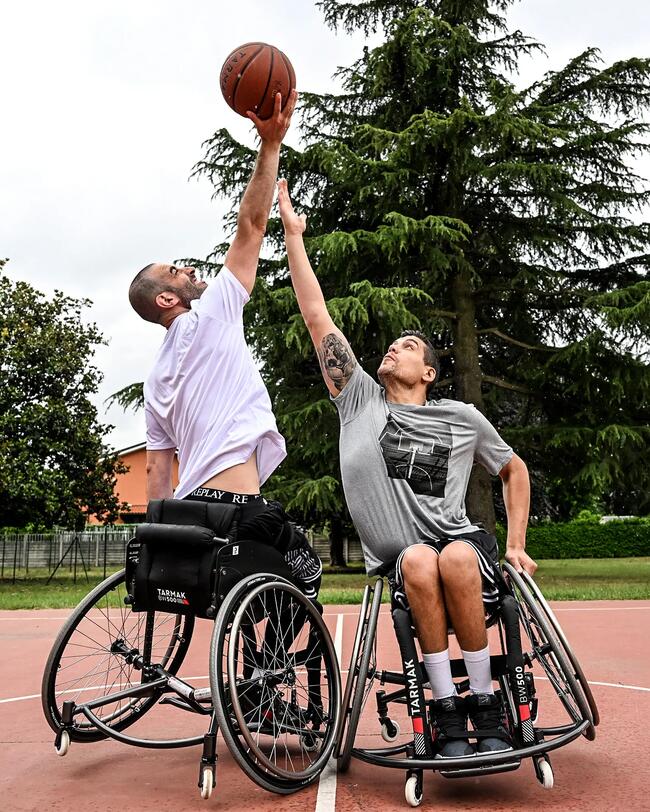 Zwei Rollstuhlbasketballspieler greifen nach dem Ball. Roter Platz, grünes Gras, große Bäume im Hintergrund.