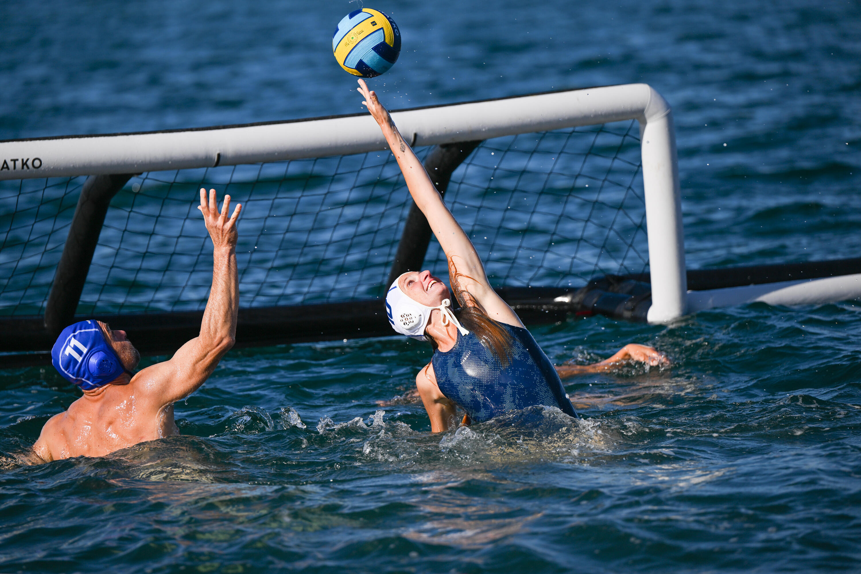 Zwei Wasserballspieler kämpfen vor einem aufblasbaren Tor im offenen Wasser um einen gelb-blauen Ball, während eine Spielerin mit weißer Kappe versucht, den Ball zu erreichen.