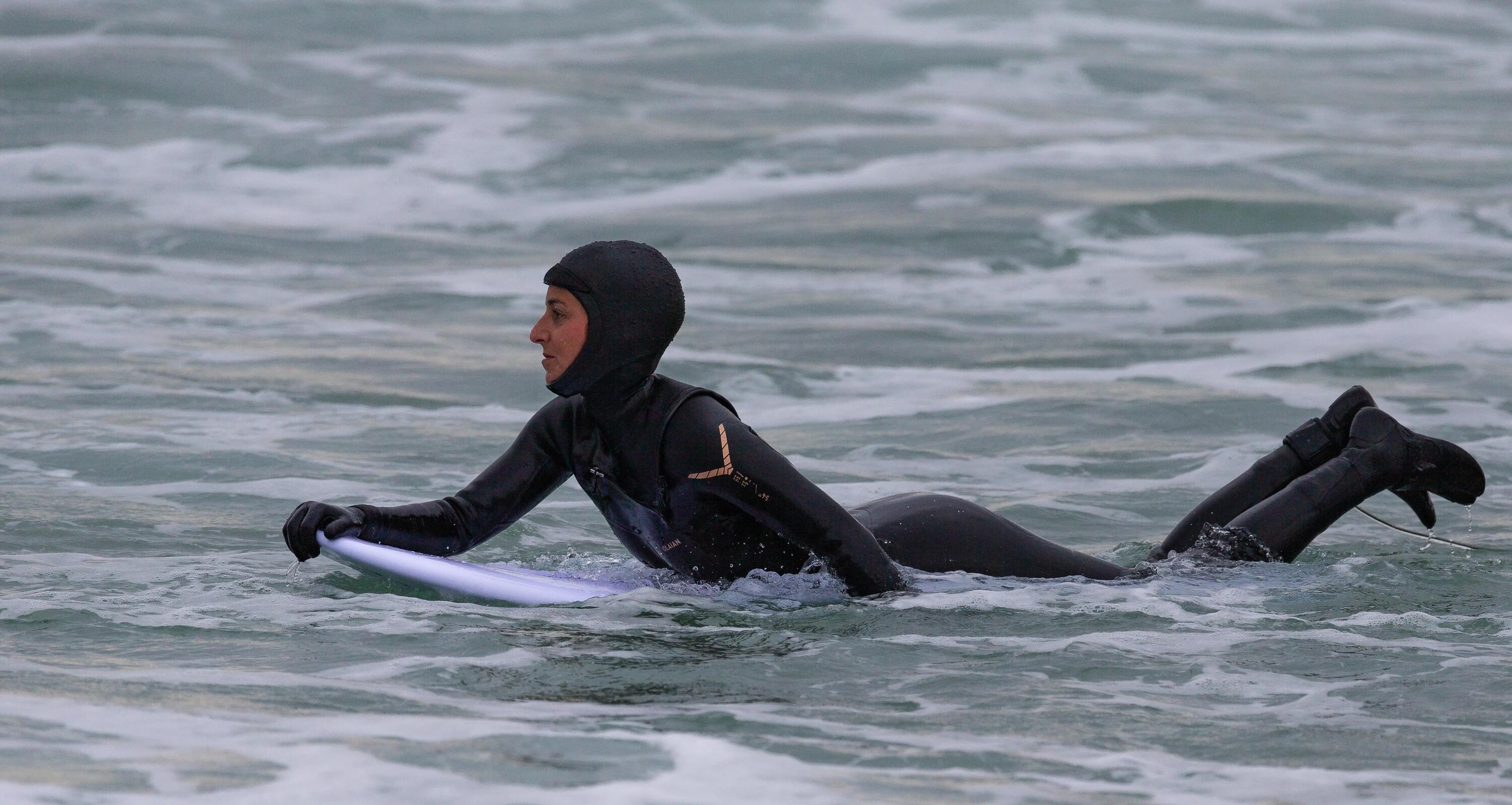 man zwemt met dik wetsuit en neopreen muts in koud water op surfboard
