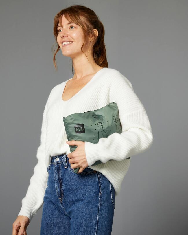 Frau in weißem Pullover mit grünem Beutel und Jeans vor grauem Hintergrund.