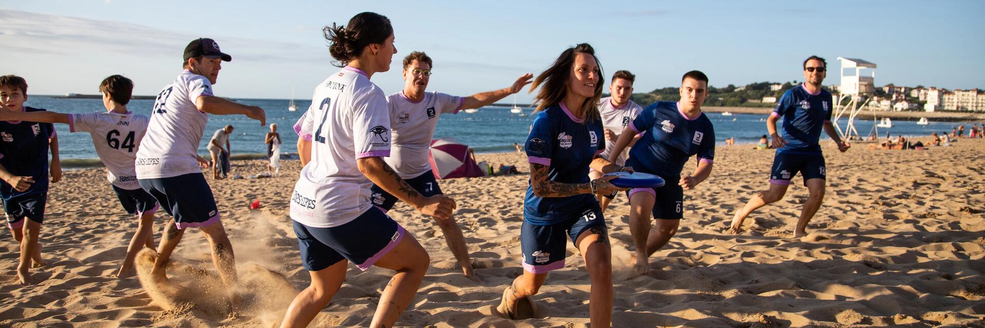 Eine Gruppe von Sportlern in Trikots spielt Ultimate Frisbee an einem weiten Sandstrand, während im Hintergrund das Meer und die Küstenlinie unter blauem Himmel zu sehen sind