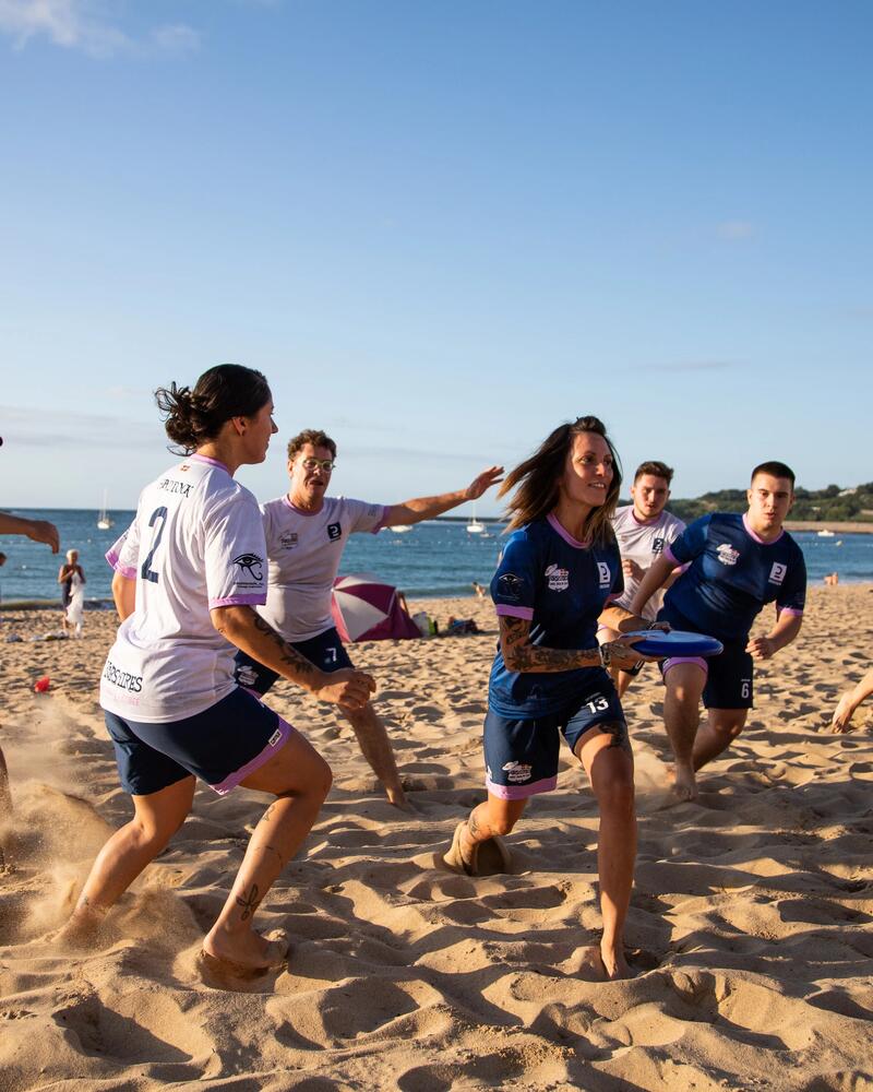 Eine Gruppe von Sportlern in Trikots spielt Ultimate Frisbee an einem weiten Sandstrand, während im Hintergrund das Meer und die Küstenlinie unter blauem Himmel zu sehen sind.