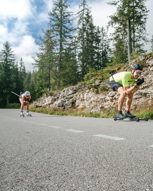 Zwei Menschen in sommerlicher Kleidung fahren in gebückter Haltung auf der Straße Rollerski.