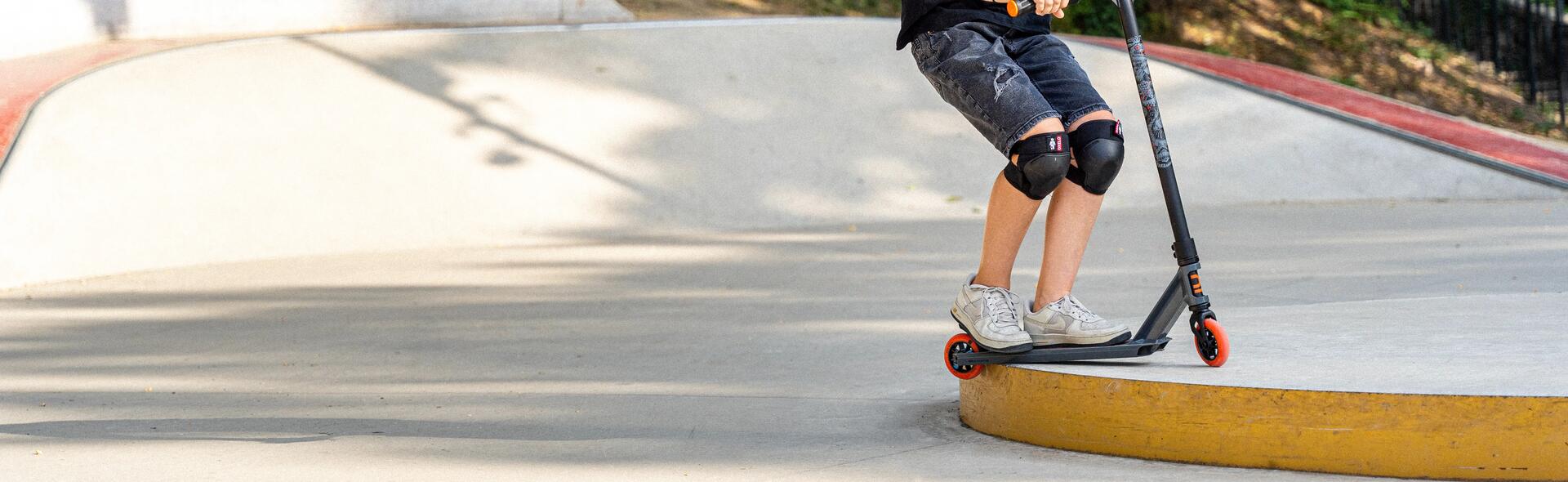 chłopiec w kasku i ochraniaczach do jazdy na hulajnodze wykonujący trik na skateparku