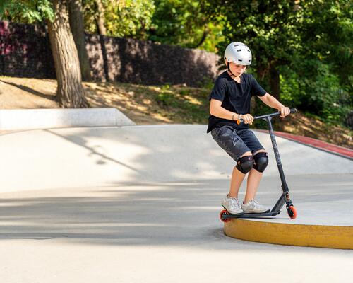 chłopiec w kasku i ochraniaczach do jazdy na hulajnodze wykonujący trik na skateparku