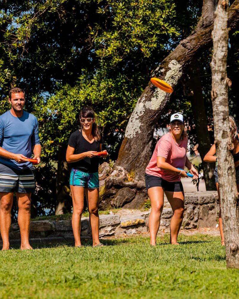 Eine Gruppe junger Erwachsener spielt auf einer Wiese im Schatten von Bäumen Discgolf oder Ultimate Frisbee; eine orangefarbene Wurfscheibe fliegt gerade durch die Luft.