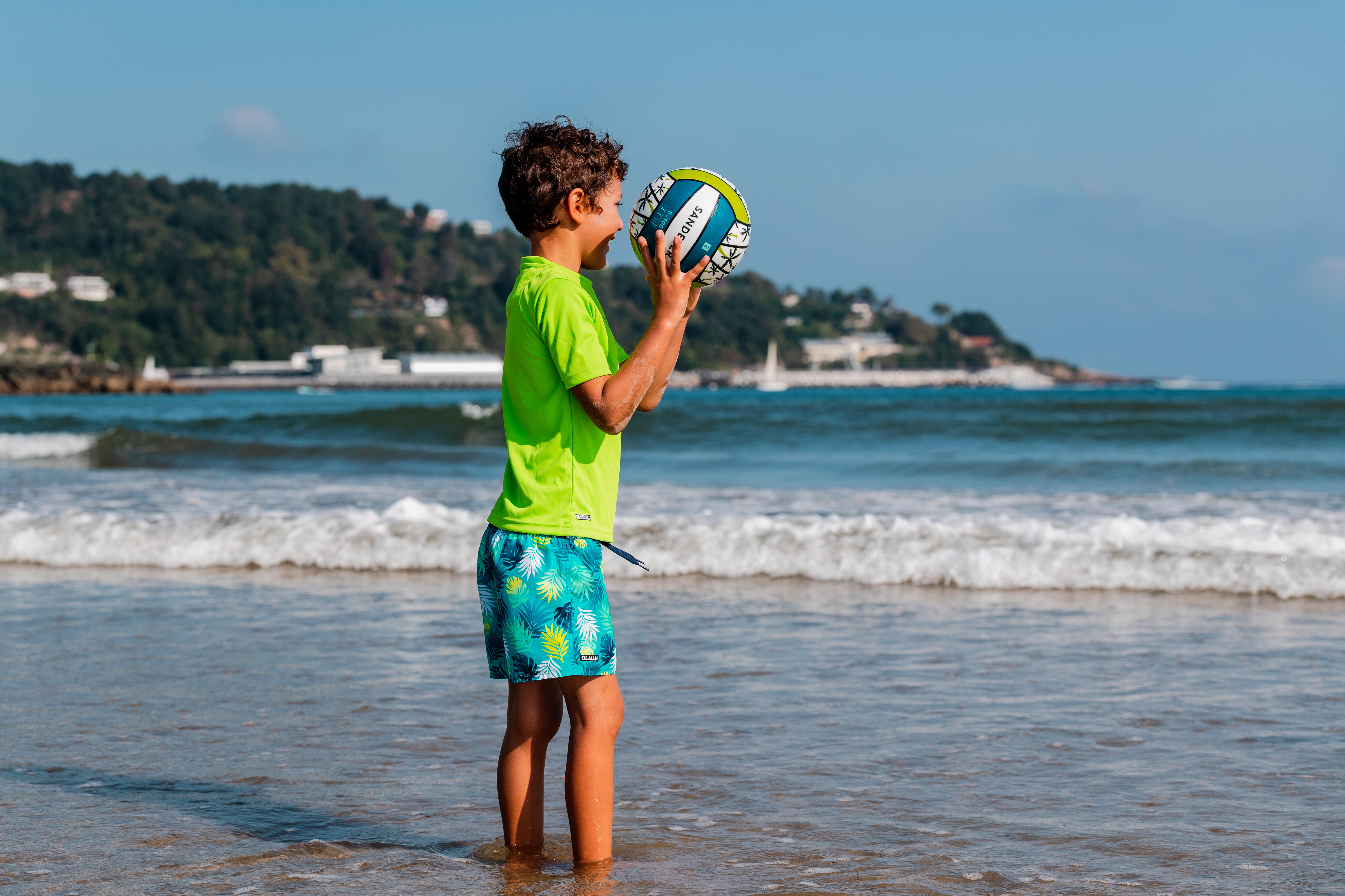 Child holding a beach volleyball while standing in shallow ocean water.