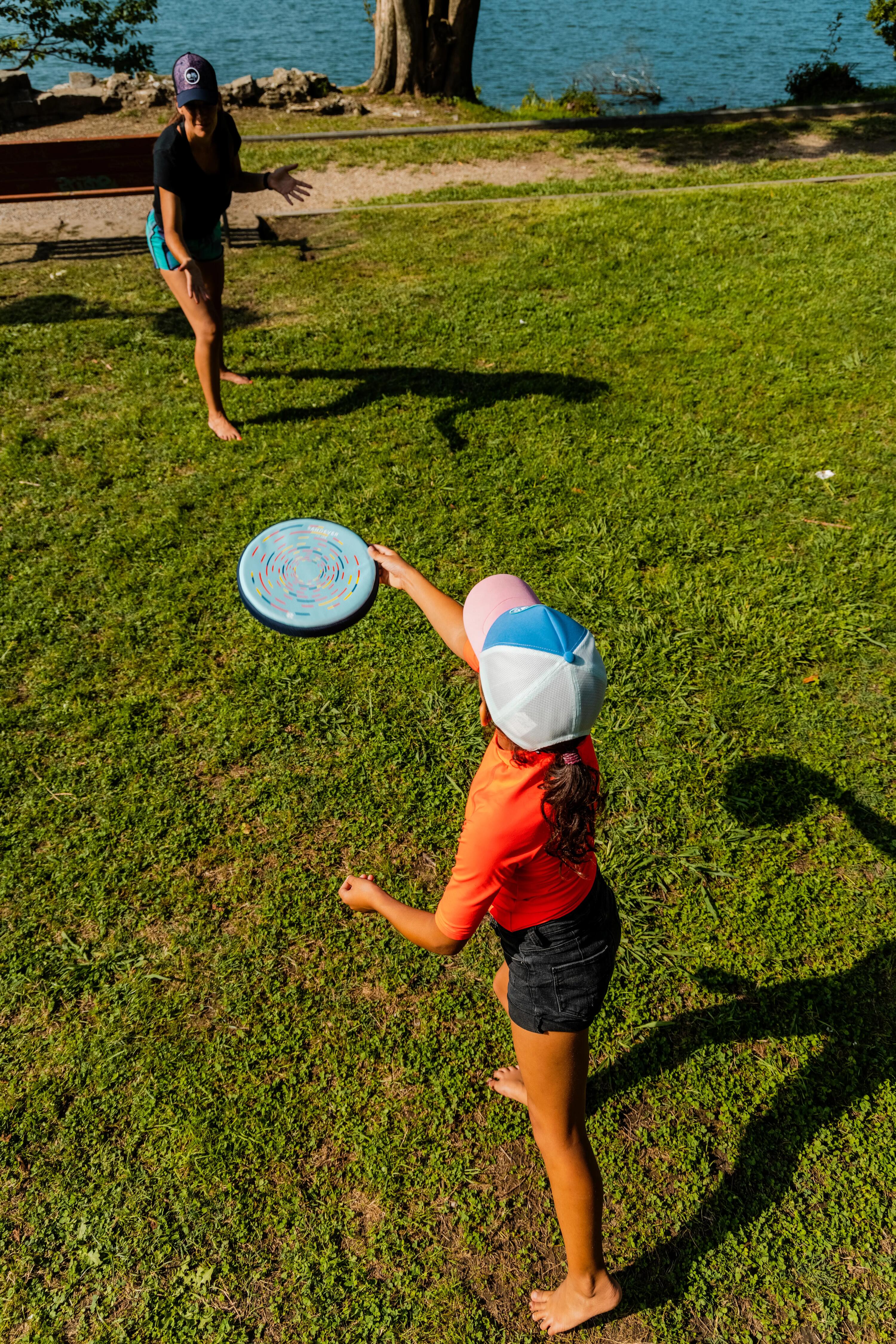 Ein Kind im orangenen Shirt und mit Kappe wirft eine blau-musternde Frisbee-Scheibe zu einer Frau auf einer grünen Wiese am Seeufer.