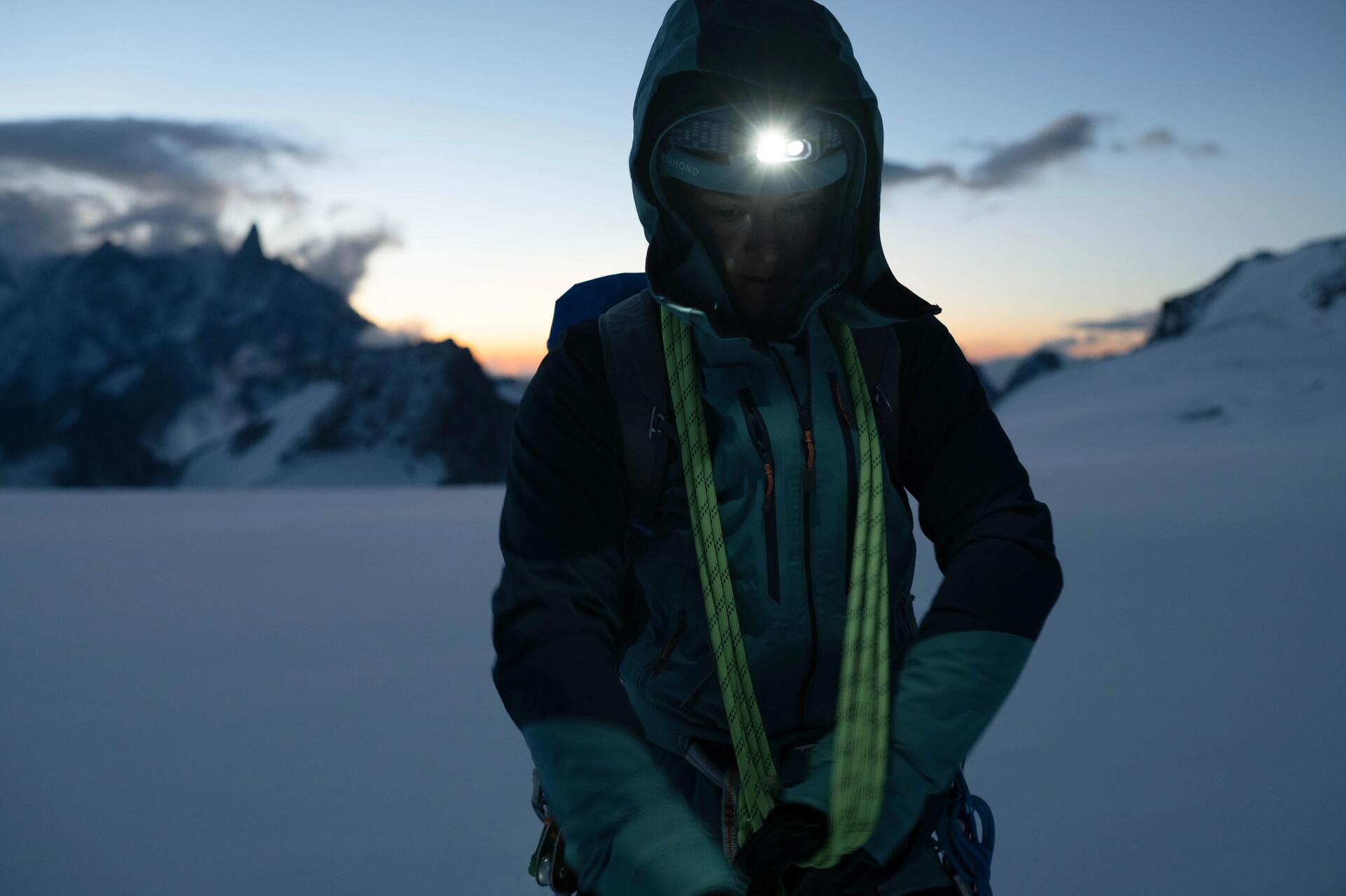 Kobieta w odzieży alpinistycznej z latarką czołową na głowie stojąca w ośnieżonych górach