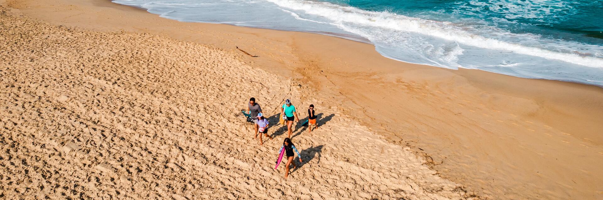 Rodzina idąca latem po plaży niosąc sprzęt i torby z przygotowanym jedzeniem na plażę