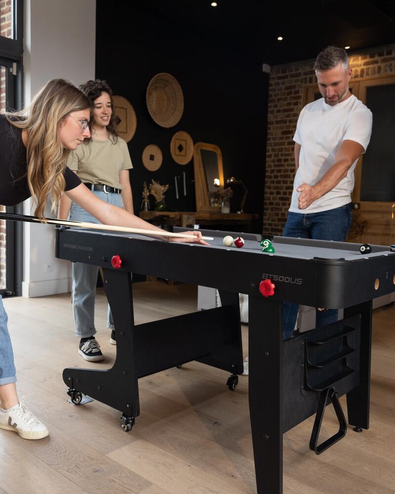 Vier junge Erwachsene spielen Billard an einem kompakten, schwarzen Multifunktionstisch in einem modern eingerichteten Wohnzimmer; eine junge Frau setzt gerade konzentriert zum Stoß an.
