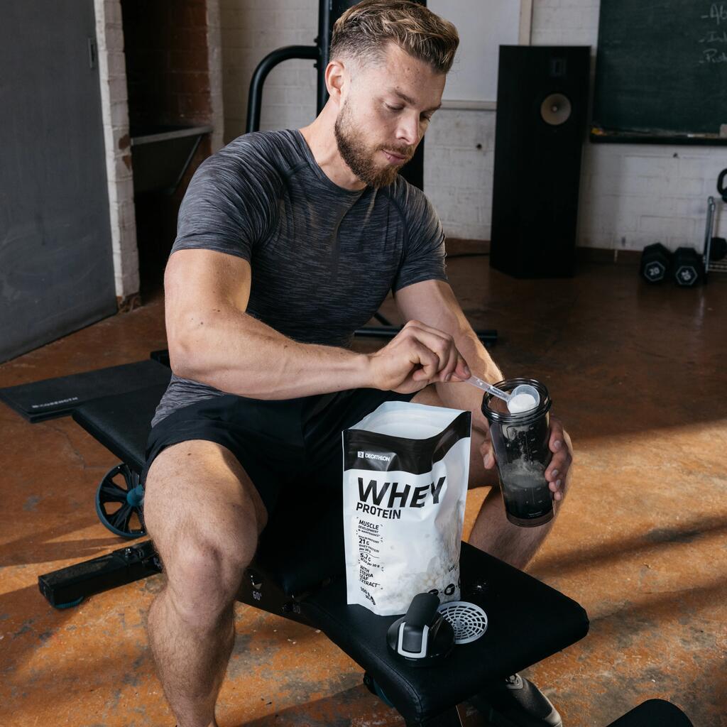 Sportaš u sivoj majici i crnom šortsu sjedi na klupi u teretani i mjericom dodaje Whey protein u prahu iz vrećice u shaker za pripremu napitka.