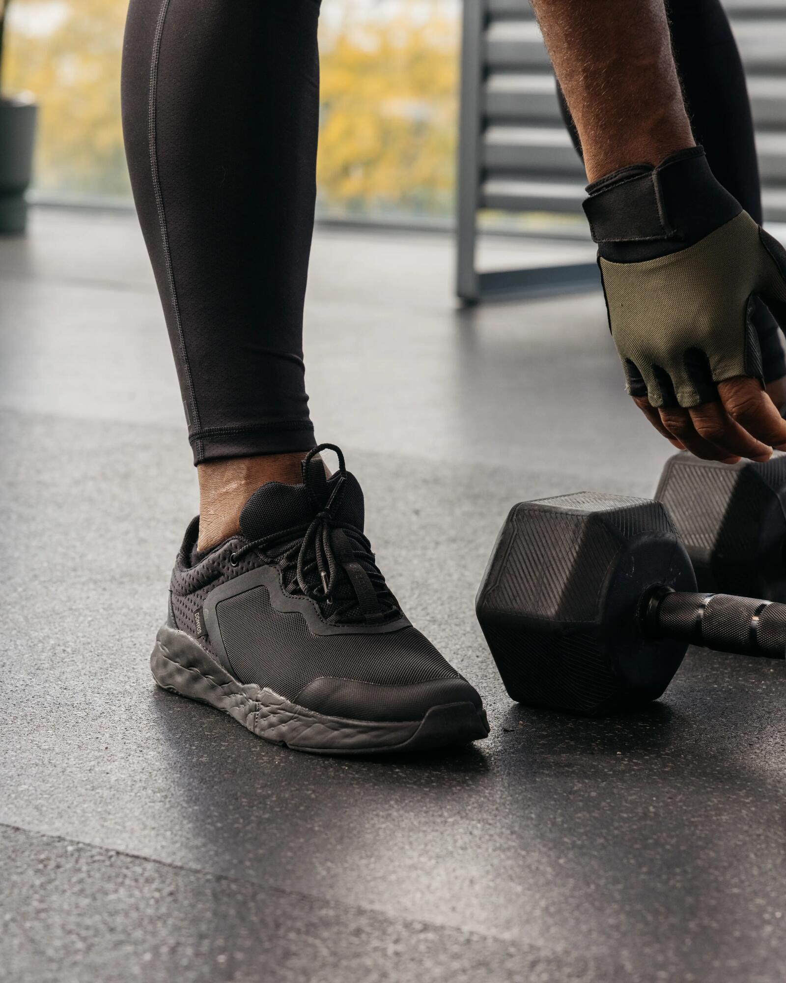 Eine Person in Sportkleidung und Fitnesshandschuhen beugt sich in einem Fitnessstudio zu zwei Kurzhanteln herab.