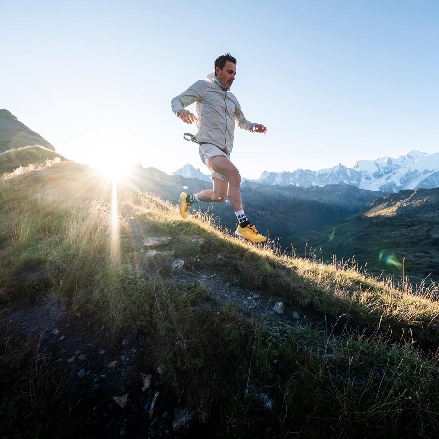 Mann läuft bei Sonnenaufgang in heller Kleidung und gelben Schuhen einen grasbewachsenen Hang mit Bergen im Hintergrund hinauf.