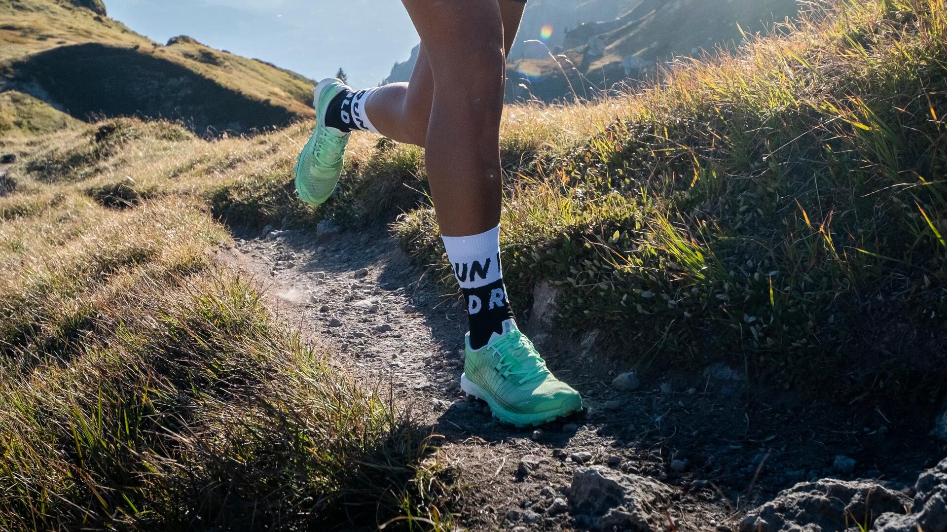 Läufer mit mintgrünen Schuhen auf einem schmalen Bergpfad, helle Sonnenstrahlen, grüne und braune Wiesen, felsige Landschaft