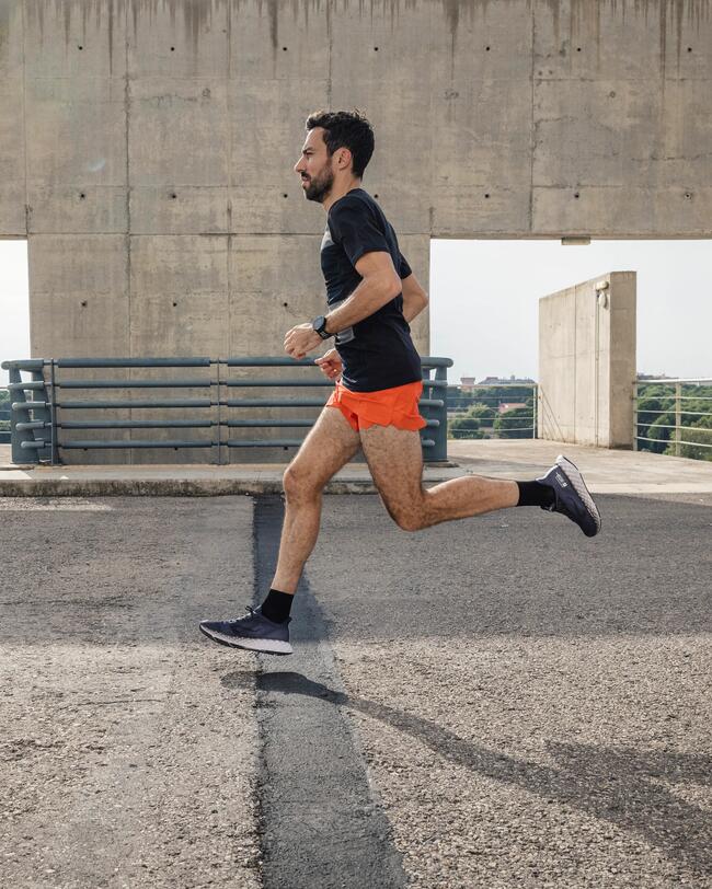 Mann in schwarzen Schuhen und oranger Sporthose läuft über grauen Asphalt vor Betonwänden mit rechteckigen Öffnungen.