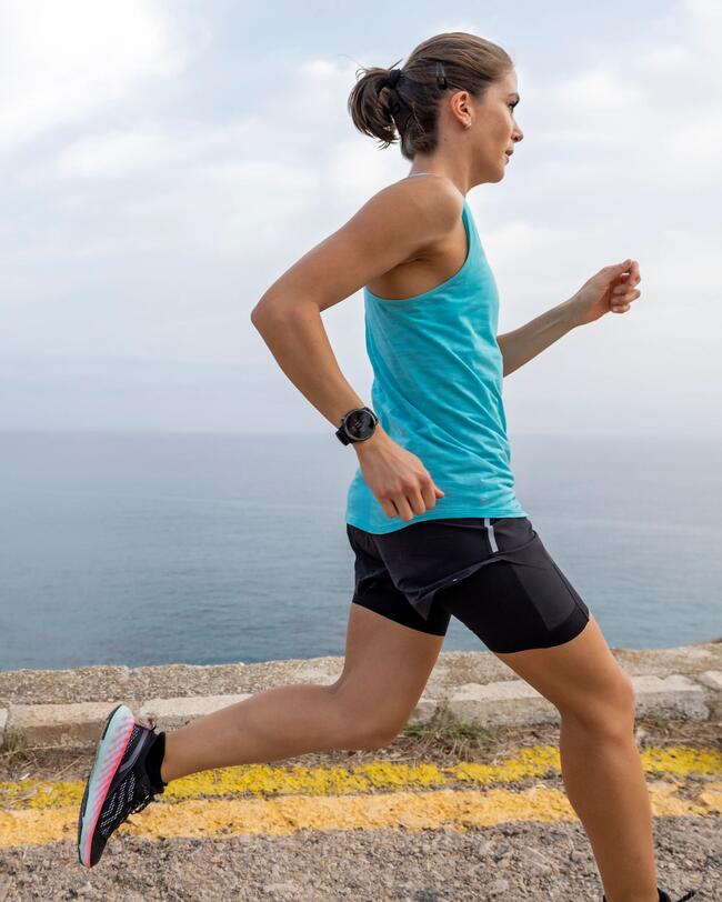 Frau joggt an Felsküste entlang, blaues Top, schwarze Shorts, grauer Himmel, blaues Meer.