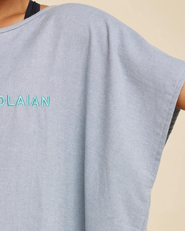  Eine Person, von hinten und leicht seitlich aufgenommen, trägt einen grauen Mikrofaser-Poncho mit aquafarbenem "OLAIAN"-Logo auf dem Rücken, der für einfache Handhabung und schnelles Überziehen steht.