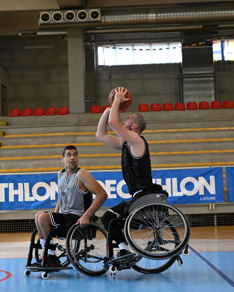 Zwei Rollstuhlbasketballspieler befinden sich auf einem Hallenspielfeld, wobei einer im Vordergrund zum Sprungwurf ansetzt, während der andere ihn aufmerksam beobachtet.