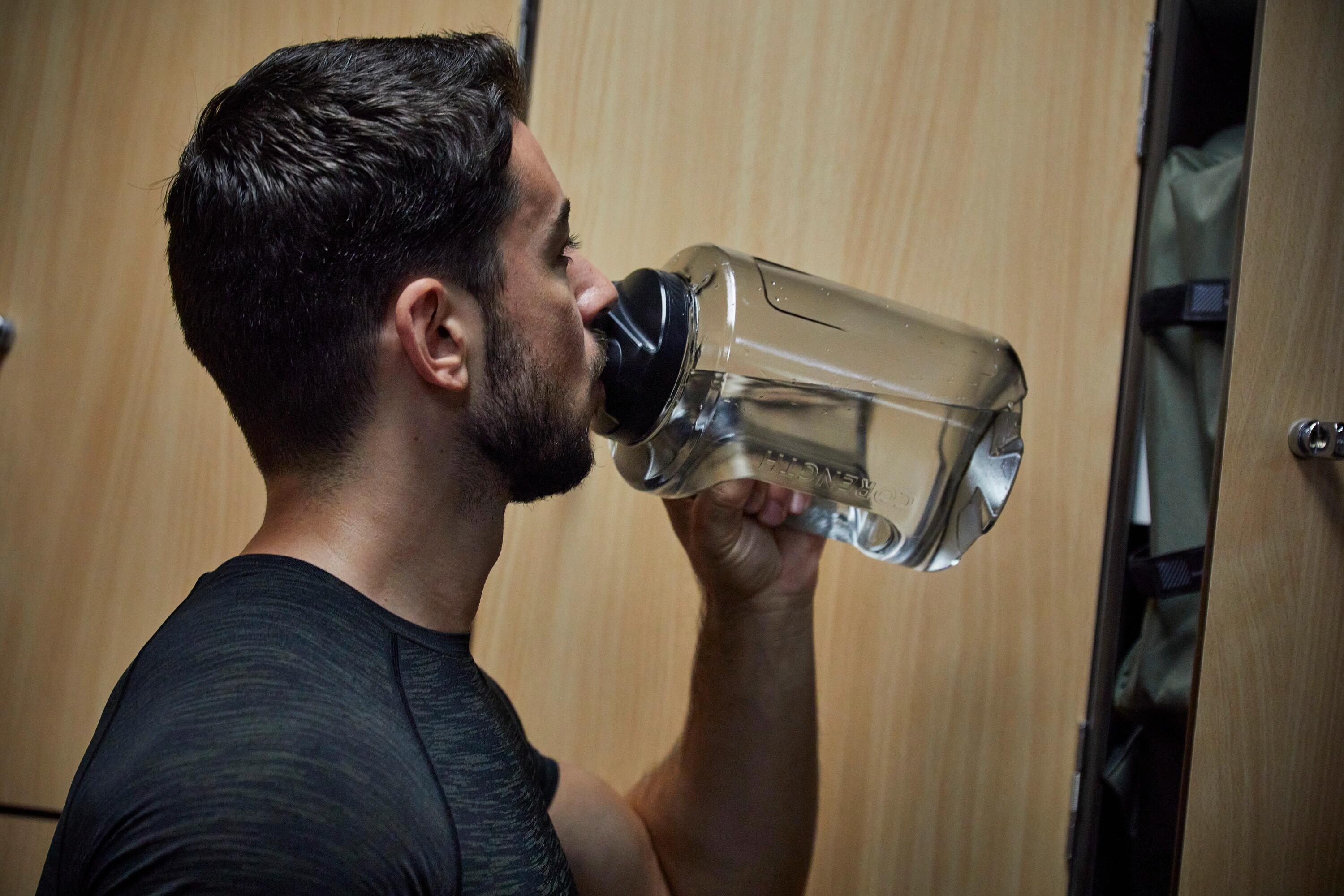 Mann trinkt Wasser aus einer großen, transparenten Gallonen-Trinkflasche in einer Umkleidekabine.