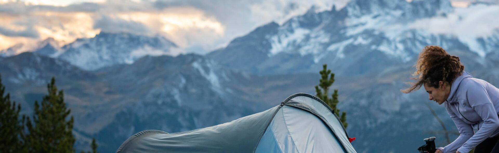 kobieta trzymająca plecak turystyczny i kije trekkingowe przed namiotem  rozłożonym w plenerze