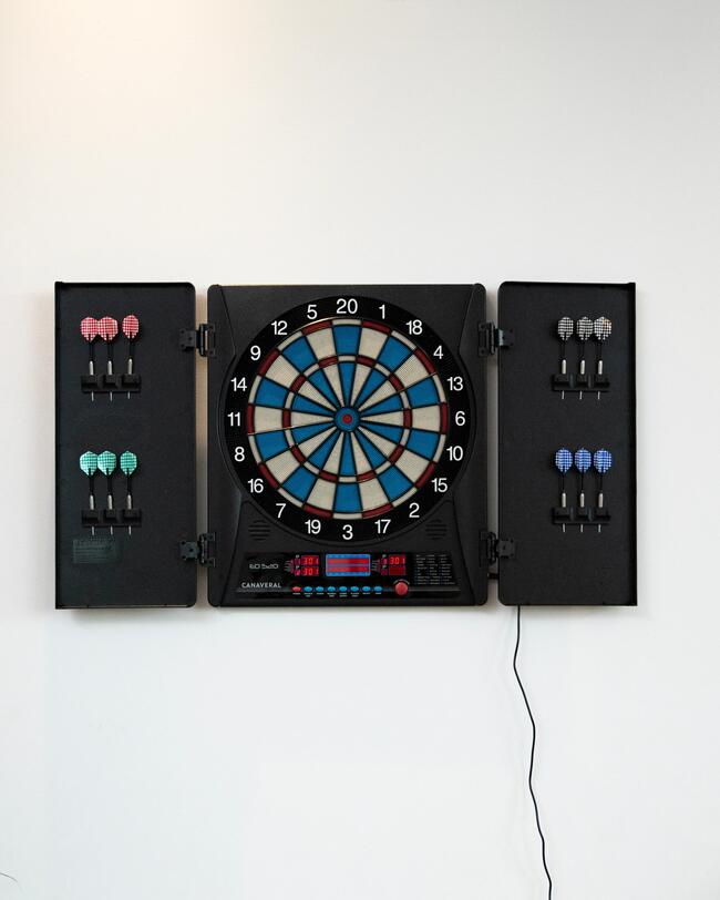 Auf einem weißen Hintergrund ist eine schwarze elektronische Dartscheibe zu sehen, deren Seitenflügel geöffnet sind und darin Reihen von Dartpfeilen in verschiedenen Farben präsentieren.