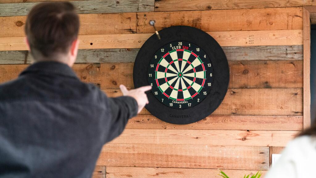 Ein Mann von hinten wirft gerade einen Pfeil auf eine Dartscheibe, die an einer Holzverkleidung hängt.