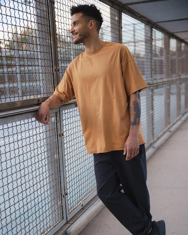Man lehnt lässig an einem Gitter. Er trägt eine schwarze Jpgginghose und ein orangenes T-Shirt