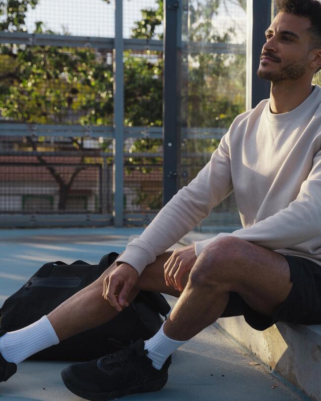  Ein Mann mit einem beigefarbenen Sweatshirt und kurzen Hosen sitzt draußen, lehnt sich zurück und blickt nach oben, während Sonnenlicht von rechts auf sein Gesicht fällt.