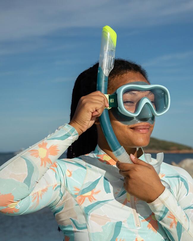 Frau steht in der Sonne, schiebt Schnorchel in Riemen der Schnorchelmaske, trägt UV-Neopren-Shirt