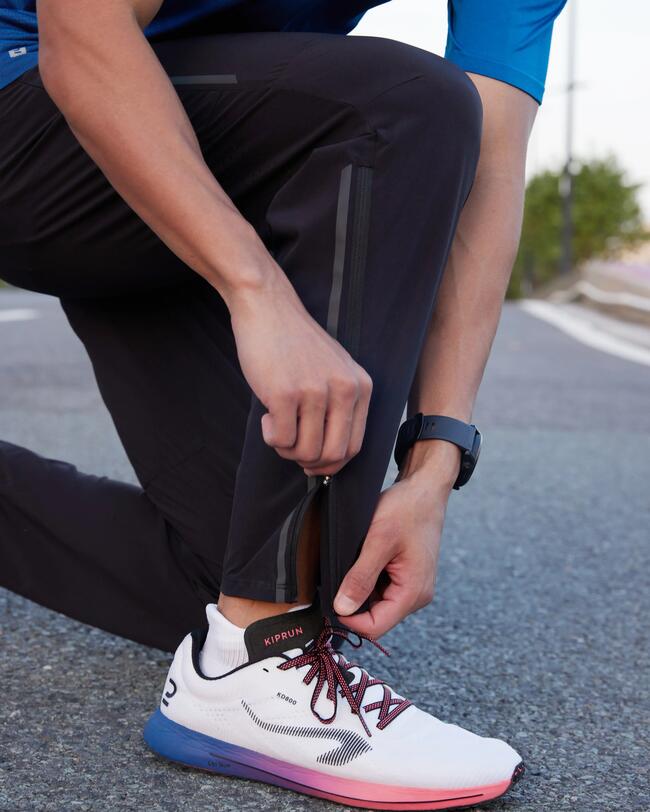 Person in blauem Hemd und schwarzen Hosen bindet weiße Laufschuhe auf grauem Asphalt.