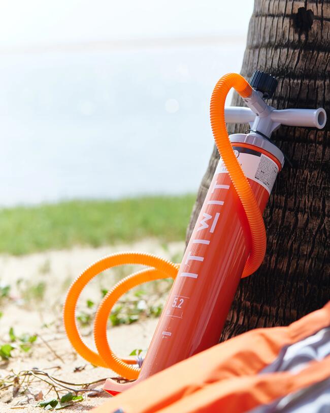 Orange Pumpe mit flexiblem Schlauch an Baum, sandiger Strand und blaues Wasser im Hintergrund.