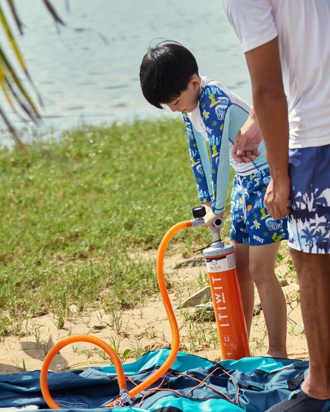 Junge in blauer Badekleidung pumpt aufblasbares Boot mit orangefarbener Pumpe auf; Strand und Wasser im Hintergrund.