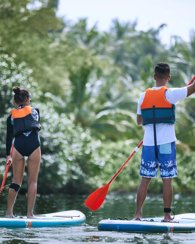 Zwei Personen auf Paddleboards mit orangefarbenen Auftriebswesten auf ruhigem Wasser, umgeben von grünen Bäumen und blauem Himmel.