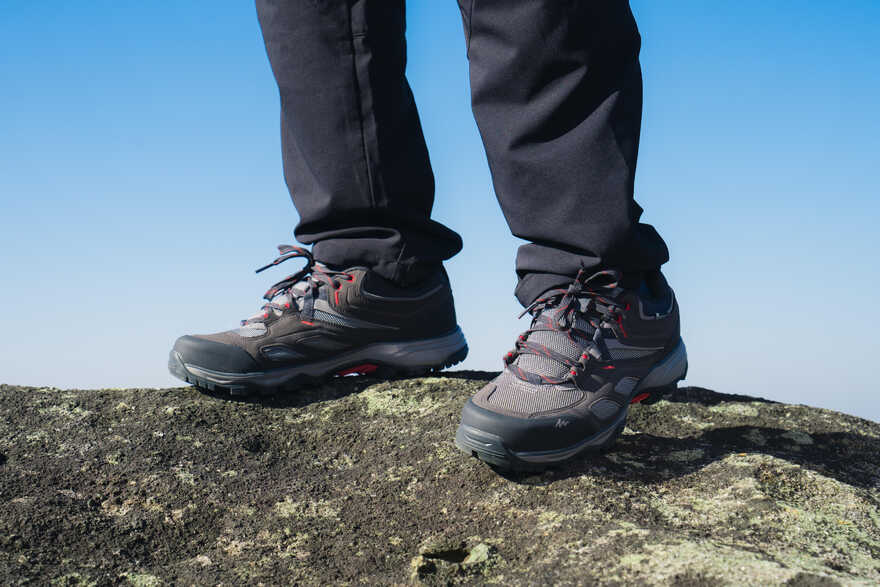 Muške noge u tamnosivim Quechua MH100 vodootpornim cipelama za planinarenje dok stoji na kamenitoj površini pod vedrim plavim nebom.