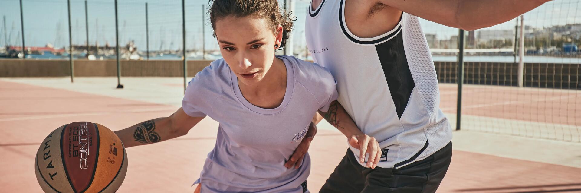 Zwei Personen spielen Basketball auf einem roten Platz bei sonnigem Wetter, dynamische Bewegung und Konzentration.