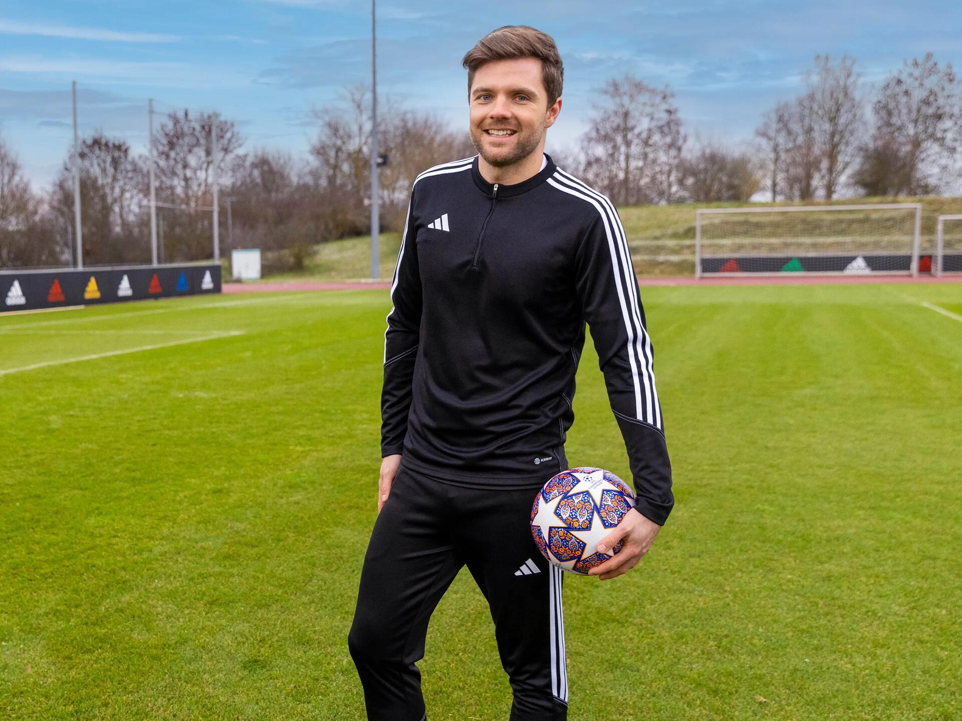 Uśmiechnięty trener piłkarski w odzieży sportowej stojący na boisku trzymając piłę do piłki nożnej