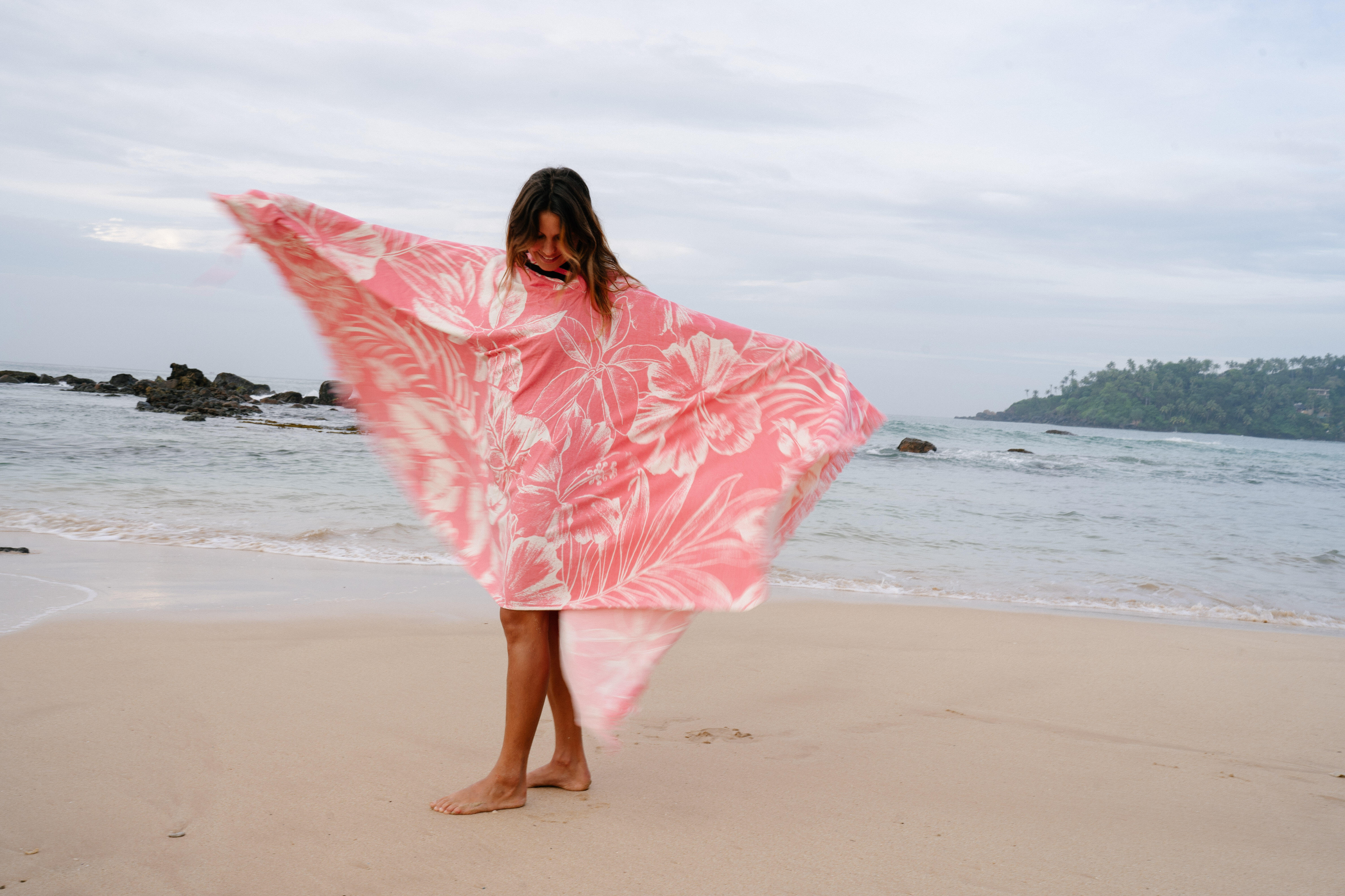 Woman wrapped in a pink floral beach towel while standing on the shoreline.