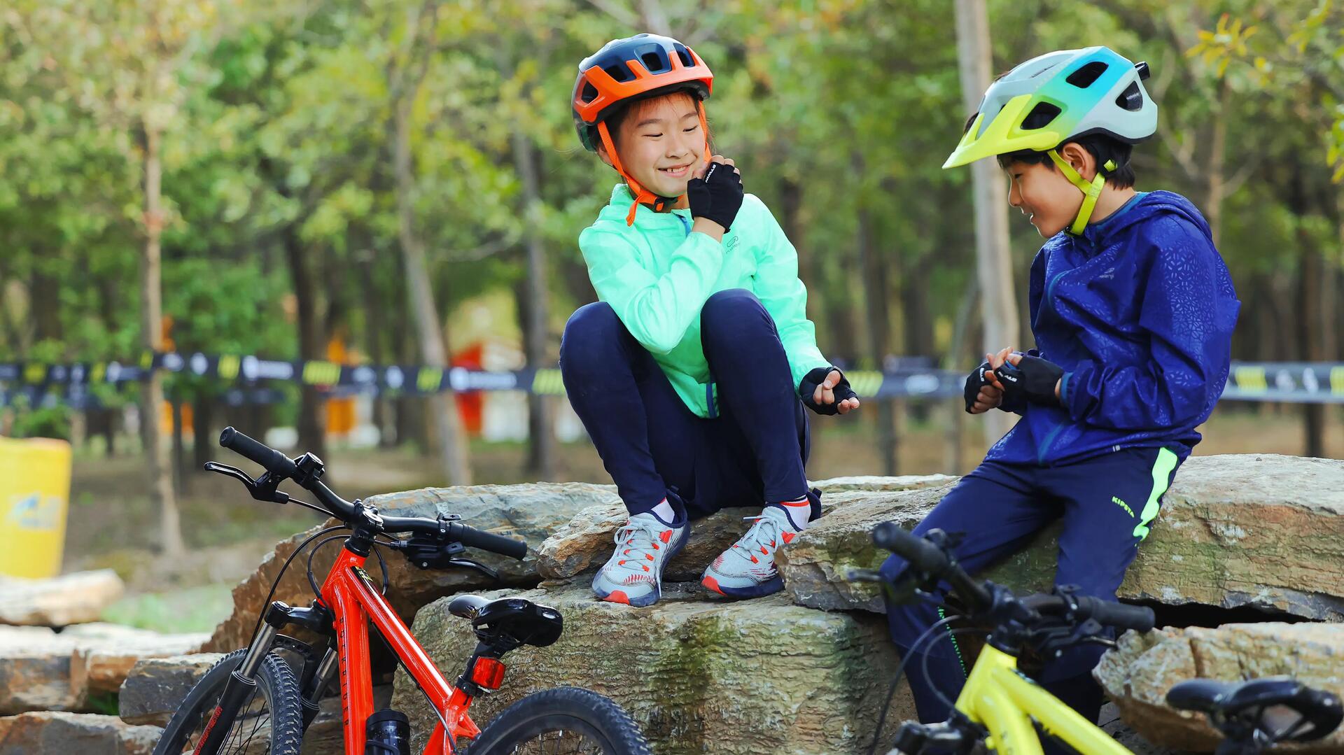 dzieci ubrane w kaski  i odzież rowerową siedzą na skałce przy rowerach 